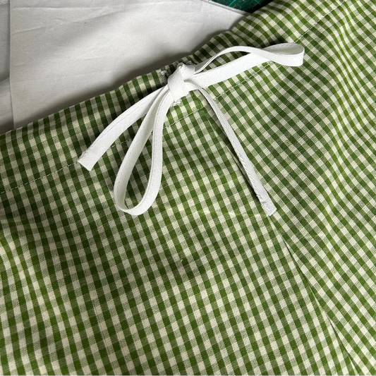 A close-up of a green and white gingham fabric item tied with a white ribbon, placed on a green cutting mat with a white fabric piece underneath.