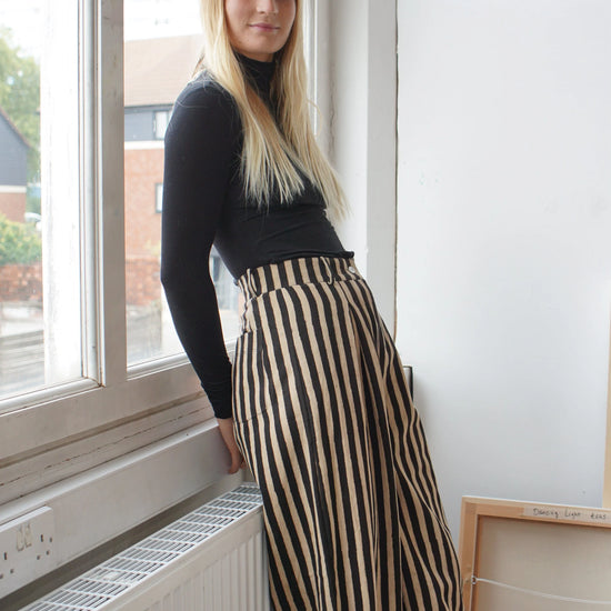 A woman leaning by a window wearing a black top and striped black and white trousers 