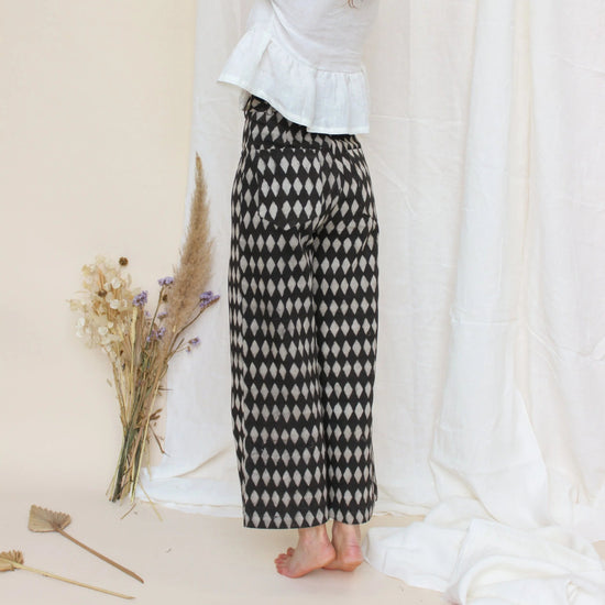 Woman standing by a white wall with heavily patterned trousers on