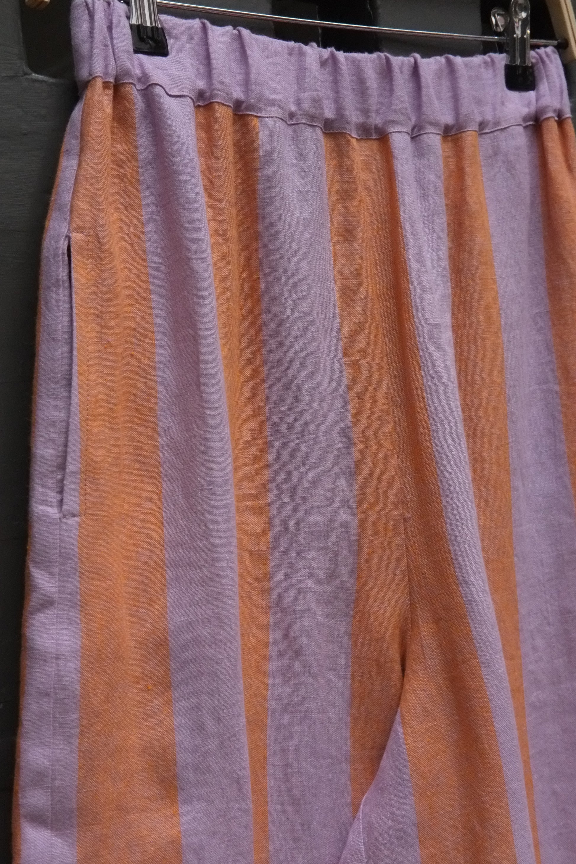 close up photo of detail of some striped linen trousers in lavender and peach showing a side pocket and elasticated waistband 