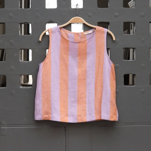 A sleeveless striped top in lavender and peach-orange hangs on a wooden hanger against a dark gray slotted wall.