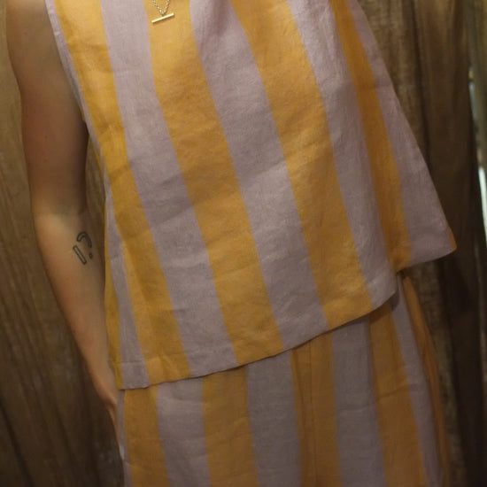 A close-up of a person wearing a sleeveless top with wide horizontal peach and lilac stripes, photographed indoors against a brown curtain.
