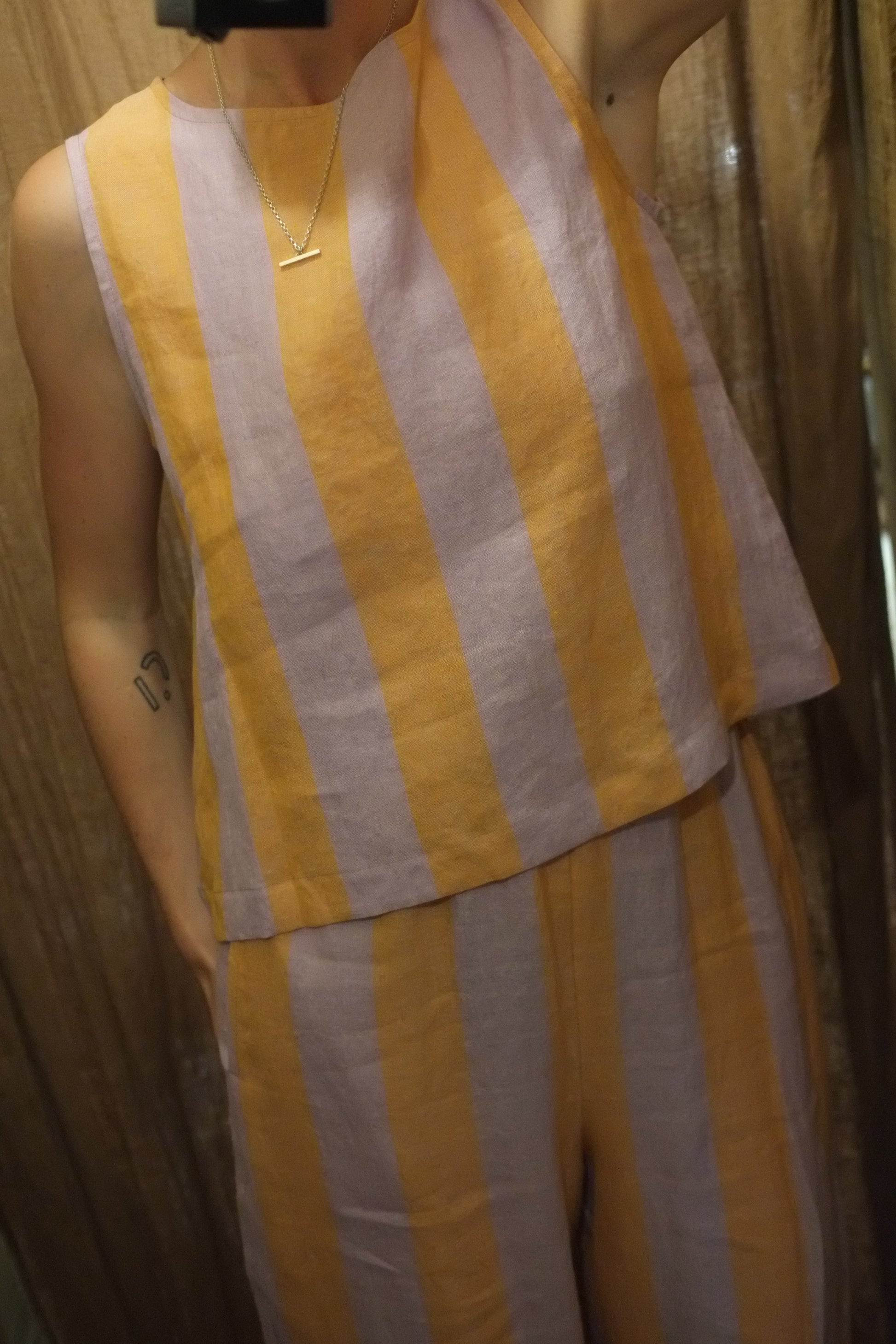 A close-up of a person wearing a sleeveless top with wide horizontal peach and lilac stripes, photographed indoors against a brown curtain.