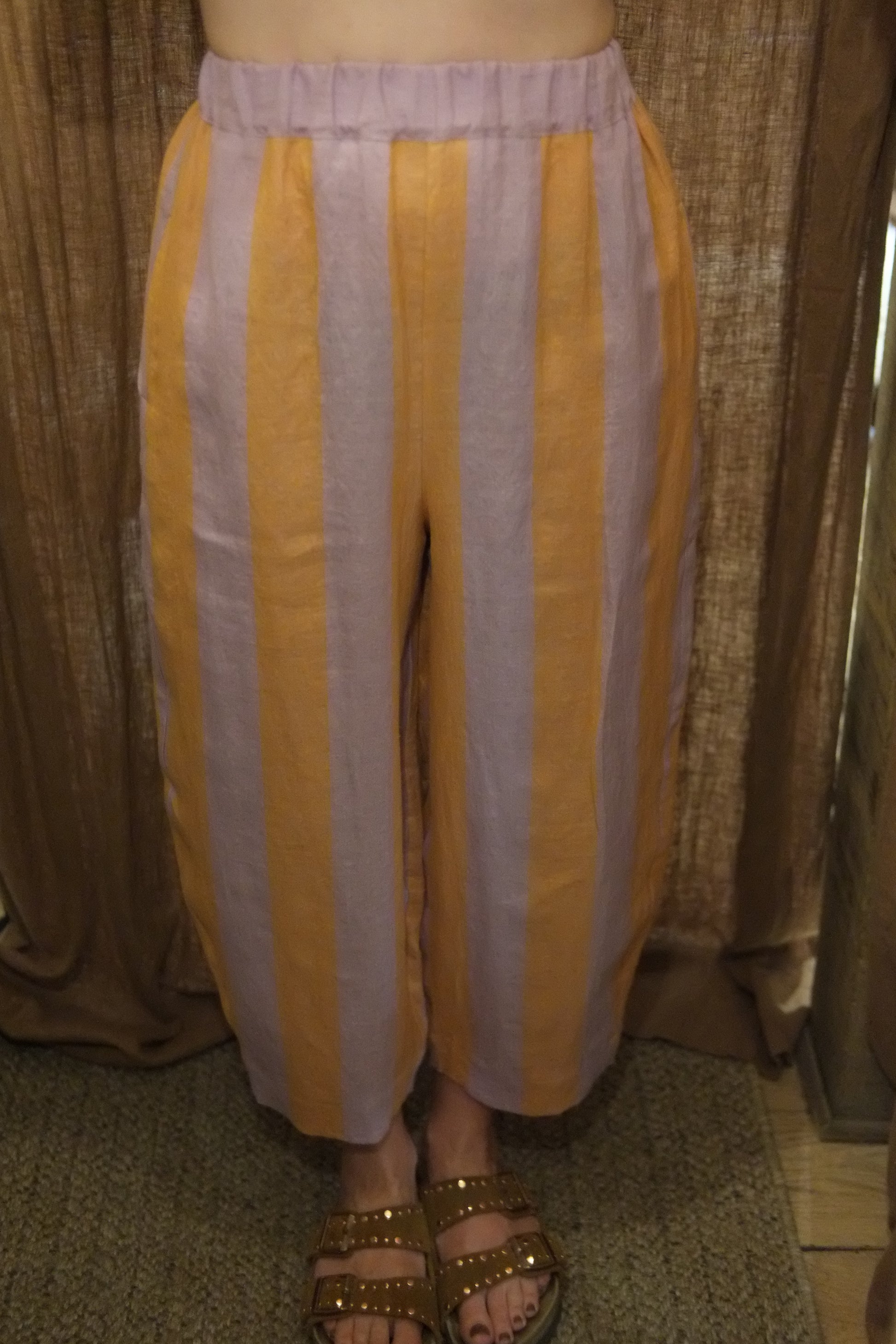 Yellow and white striped pants with a gathered elastic waistband, shown on a model wearing brown studded sandals.