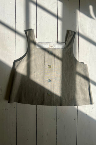 A beige linen tank top with a square neckline, thick straps, and shell buttons, displayed on a wooden surface with dappled light.