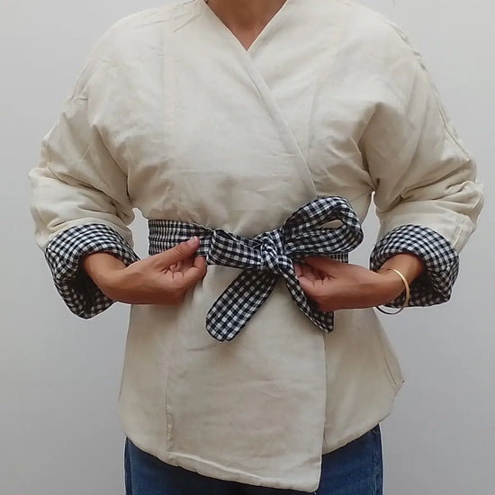 A person wearing a cream-colored wrap-style top with rolled sleeves and a matching gingham tie belt, paired with blue jeans, against a plain light grey background.