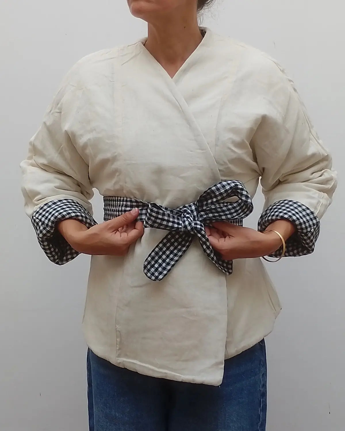 A person wearing a cream-colored wrap-style top with rolled sleeves and a matching gingham tie belt, paired with blue jeans, against a plain light grey background.