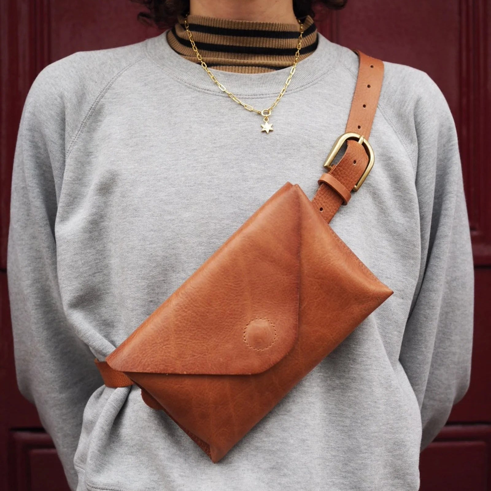 Person wearing a brown leather crossbody bag over a gray sweatshirt against a red background