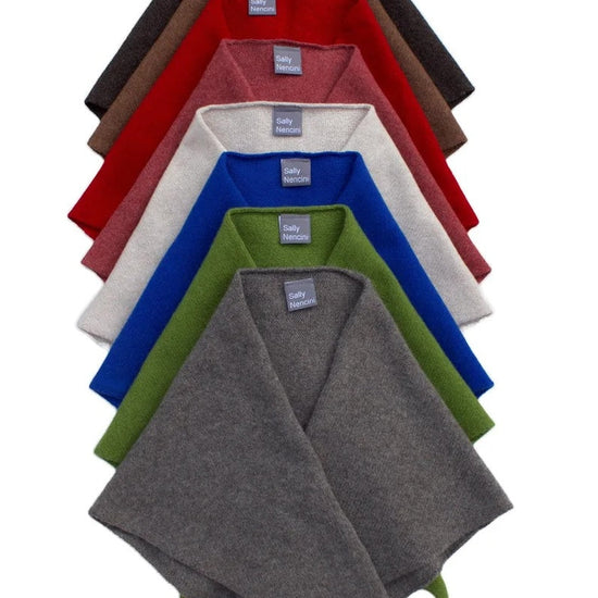 A stack of six folded knit poncho-style scarves in different colors, arranged vertically on a plain white background. The colors visible from top to bottom are dark brown, red, off-white, royal blue, olive green, and heather gray.