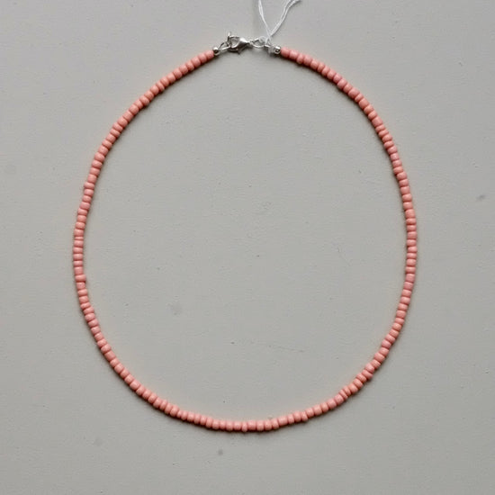 A delicate necklace made of small, uniform light pink beads arranged in a circle, featuring a silver-toned clasp at the top, displayed on a neutral gray background.