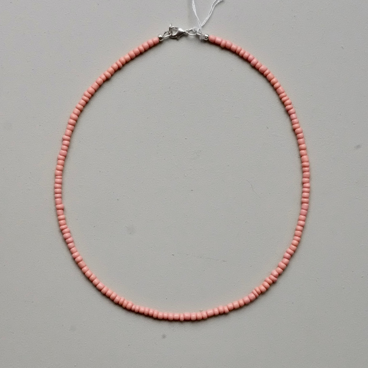A delicate necklace made of small, uniform light pink beads arranged in a circle, featuring a silver-toned clasp at the top, displayed on a neutral gray background.