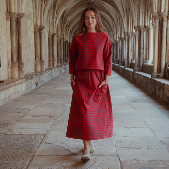A woman wearing a red  checkered two-piece set consisting of a long-sleeve top and a midi skirt, walking down a stone corridor with arches.