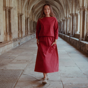 A woman wearing a red  checkered two-piece set consisting of a long-sleeve top and a midi skirt, walking down a stone corridor with arches.