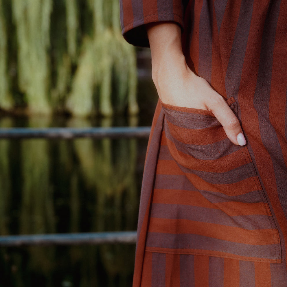 A close-up of a person wearing a rust and dark brown vertically striped dress, with one hand in a front pocket, set against a soft-focus background of a green willow tree by a body of water.