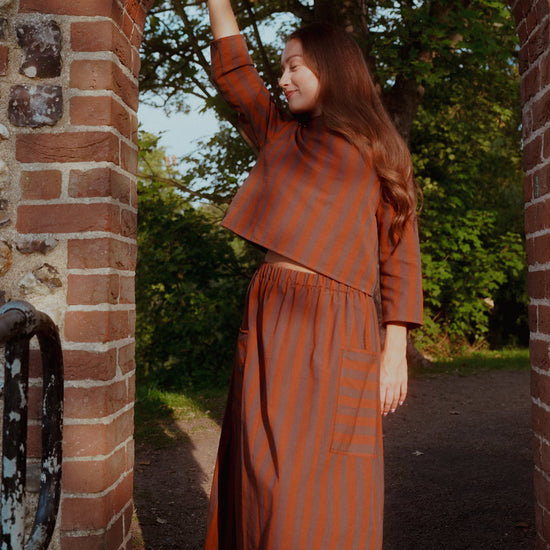A woman wearing a rust-orange and brown vertically striped two-piece set stands in a brick archway outdoors, with trees and a path visible in the background.