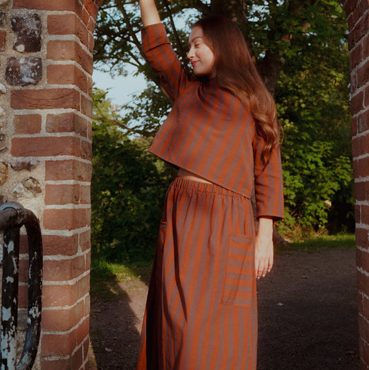 A woman wearing a rust-orange and brown vertically striped two-piece set stands in a brick archway outdoors, with trees and a path visible in the background.