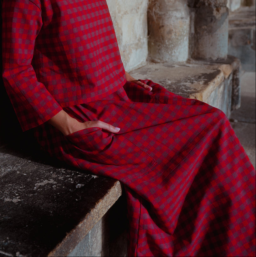 A woman wearing a red checkered long-sleeve top and skirt sits on a stone bench against a rustic stone wall.