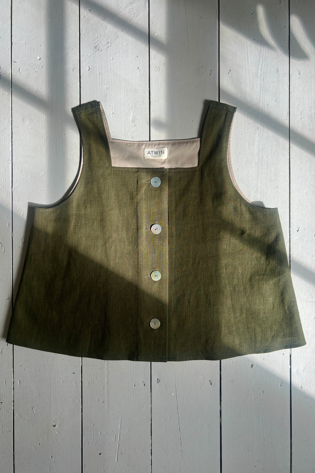 A khaki green ramie linen tank top with a square neckline, thick straps, and button-down front on a wooden background.