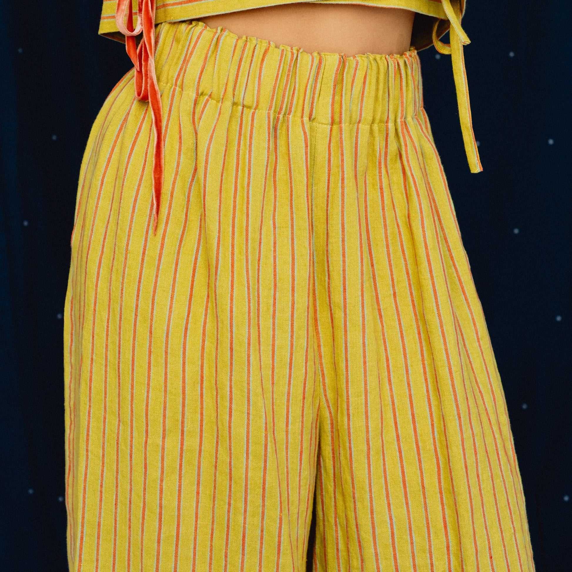 A close-up of a person wearing a yellow two-piece outfit with thin red vertical stripes, featuring a cropped wrap-style top with side ties and matching wide-leg pants, set against a dark blue starry background.