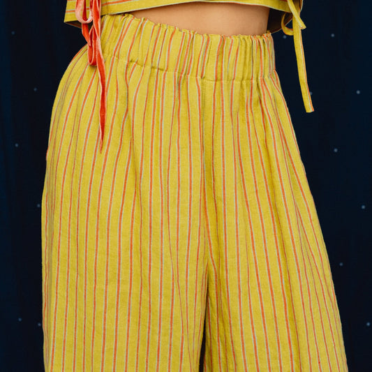 A close-up of a person wearing a yellow two-piece outfit with thin red vertical stripes, featuring a cropped wrap-style top with side ties and matching wide-leg pants, set against a dark blue starry background.