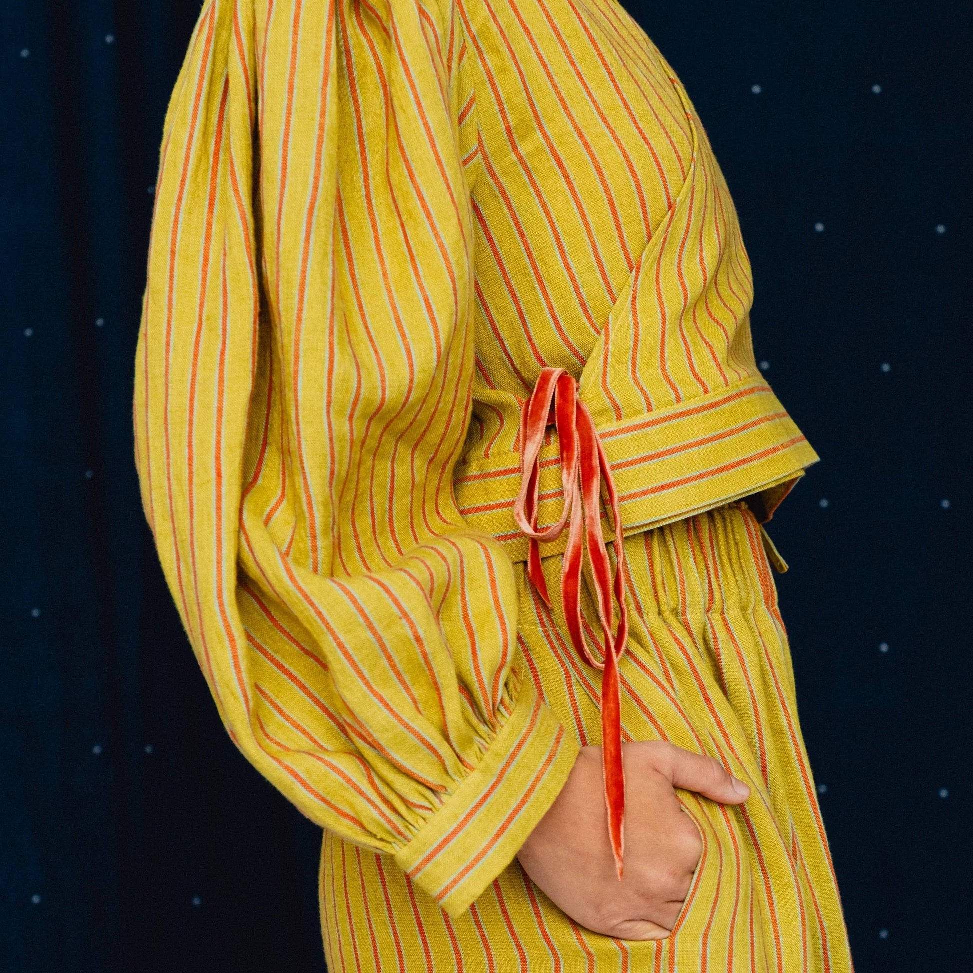 A person wearing a mustard yellow long-sleeve dress with thin red vertical pinstripes and a red waist tie, shown against a dark navy starry backdrop.