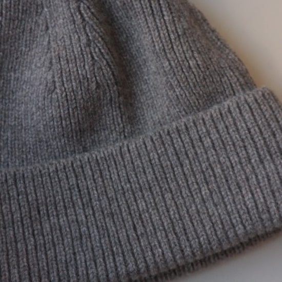 A close-up of a gray ribbed knit beanie hat, showing the texture of the fabric and folded cuff, on a light background.