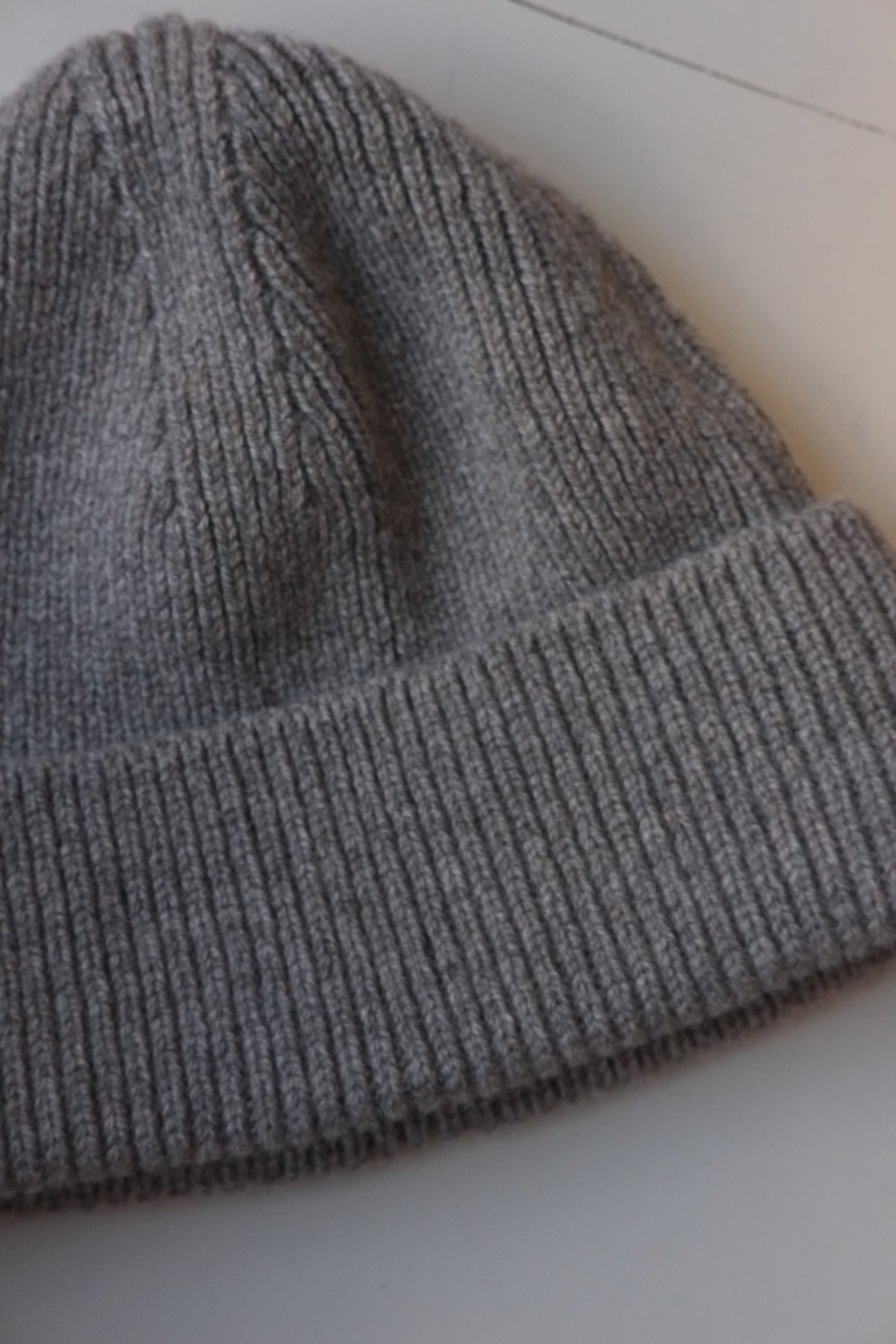 A close-up of a gray ribbed knit beanie hat, showing the texture of the fabric and folded cuff, on a light background.