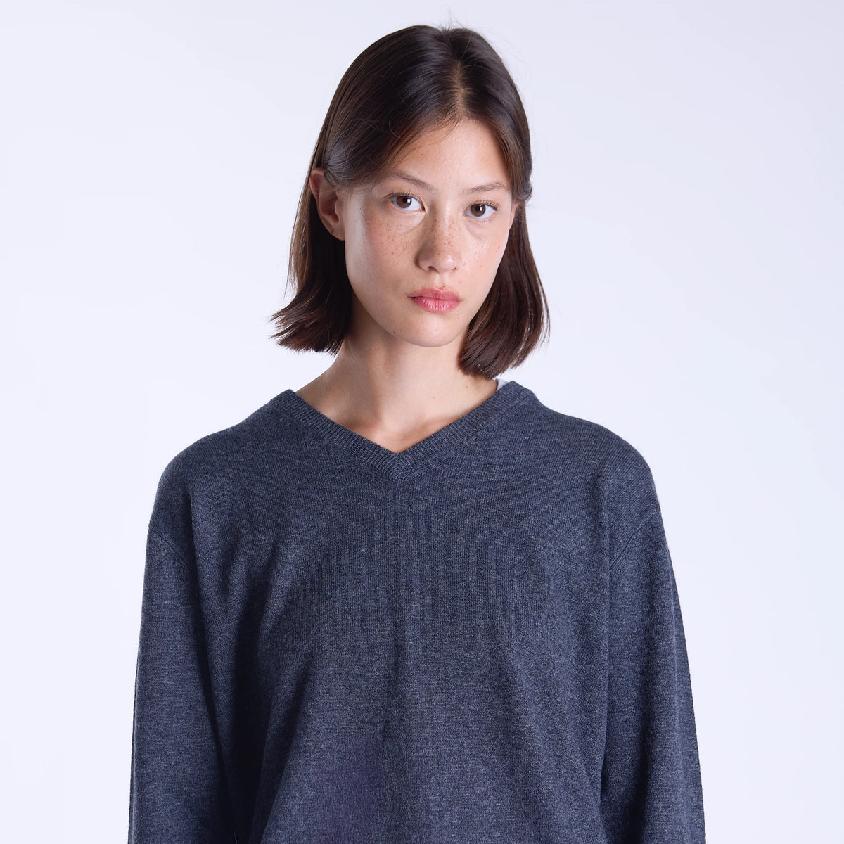 A woman modeling a dark grey long-sleeve knit sweater with a V-neck, paired with a light cream skirt, against a plain light gray studio background.