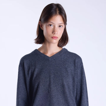 A woman modeling a dark grey long-sleeve knit sweater with a V-neck, paired with a light cream skirt, against a plain light gray studio background.