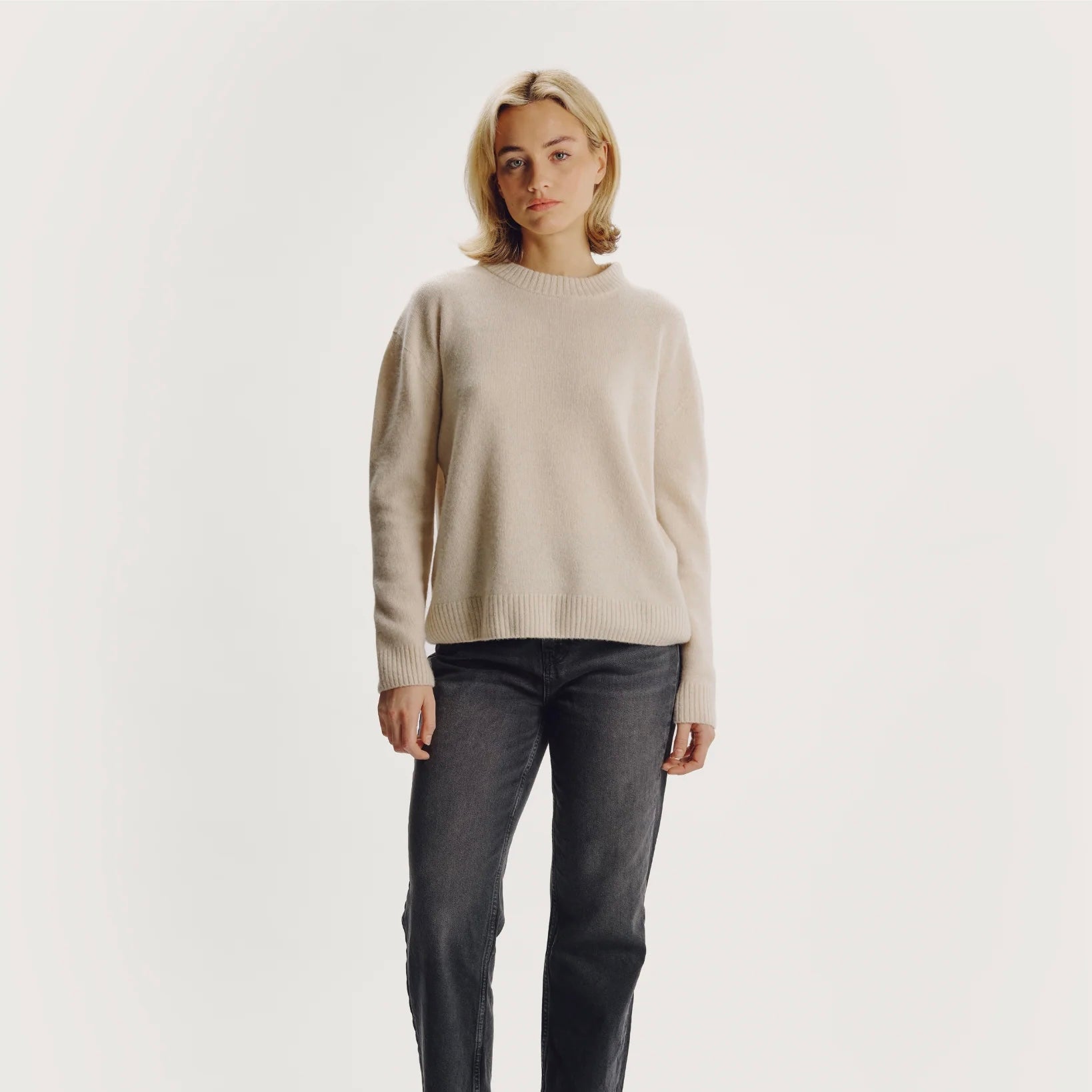 Model wearing a beige sweater and dark jeans on a plain background
