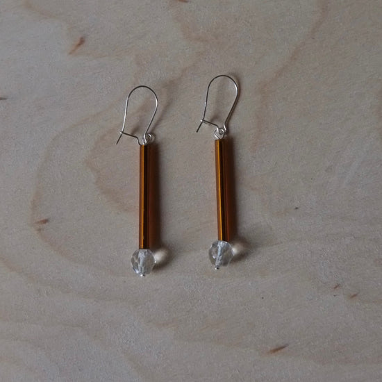 A pair of handmade dangle earrings with vertical amber-brown rods, silver fishhook wires, and small clear faceted beads at the bottom, displayed on a light wood grain surface.