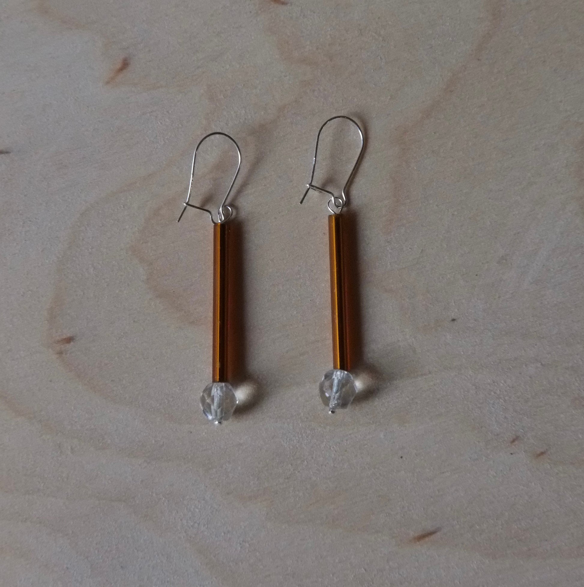 A pair of handmade dangle earrings with vertical amber-brown rods, silver fishhook wires, and small clear faceted beads at the bottom, displayed on a light wood grain surface.