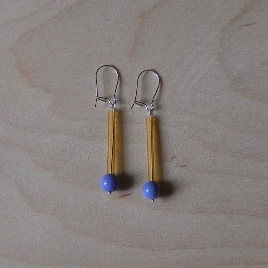 A pair of handmade dangle earrings featuring yellow strands, a small blue bead, and silver fishhook wires, displayed on a light wood grain surface.