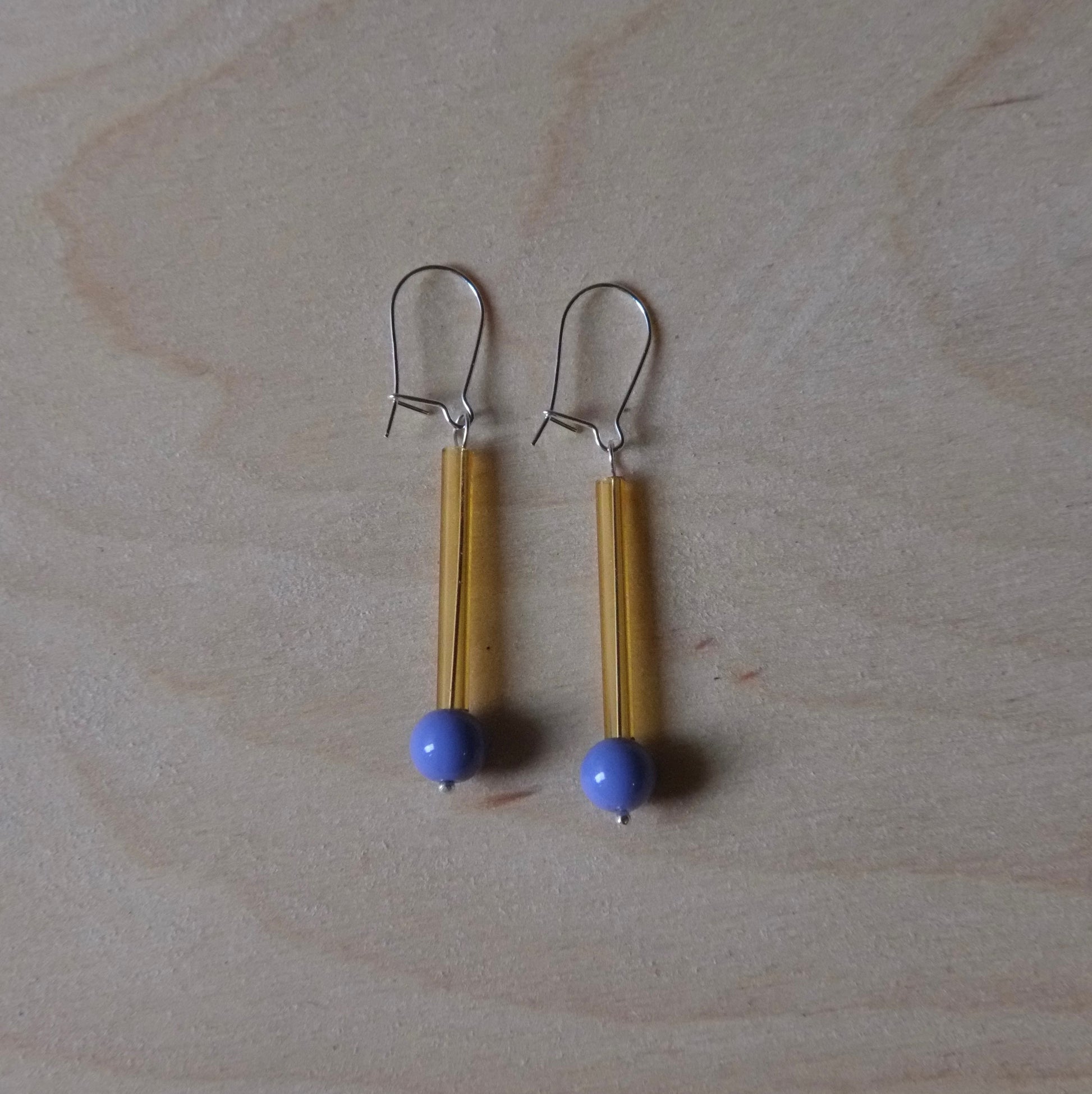 A pair of handmade dangle earrings featuring yellow strands, a small blue bead, and silver fishhook wires, displayed on a light wood grain surface.