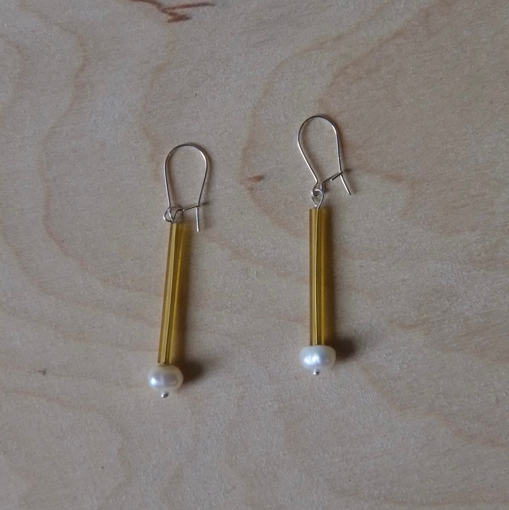 A pair of handmade dangle earrings with gold-toned metal tubes, silver fishhook backings, and small white pearl beads at the bottom, displayed on a light wood grain surface.