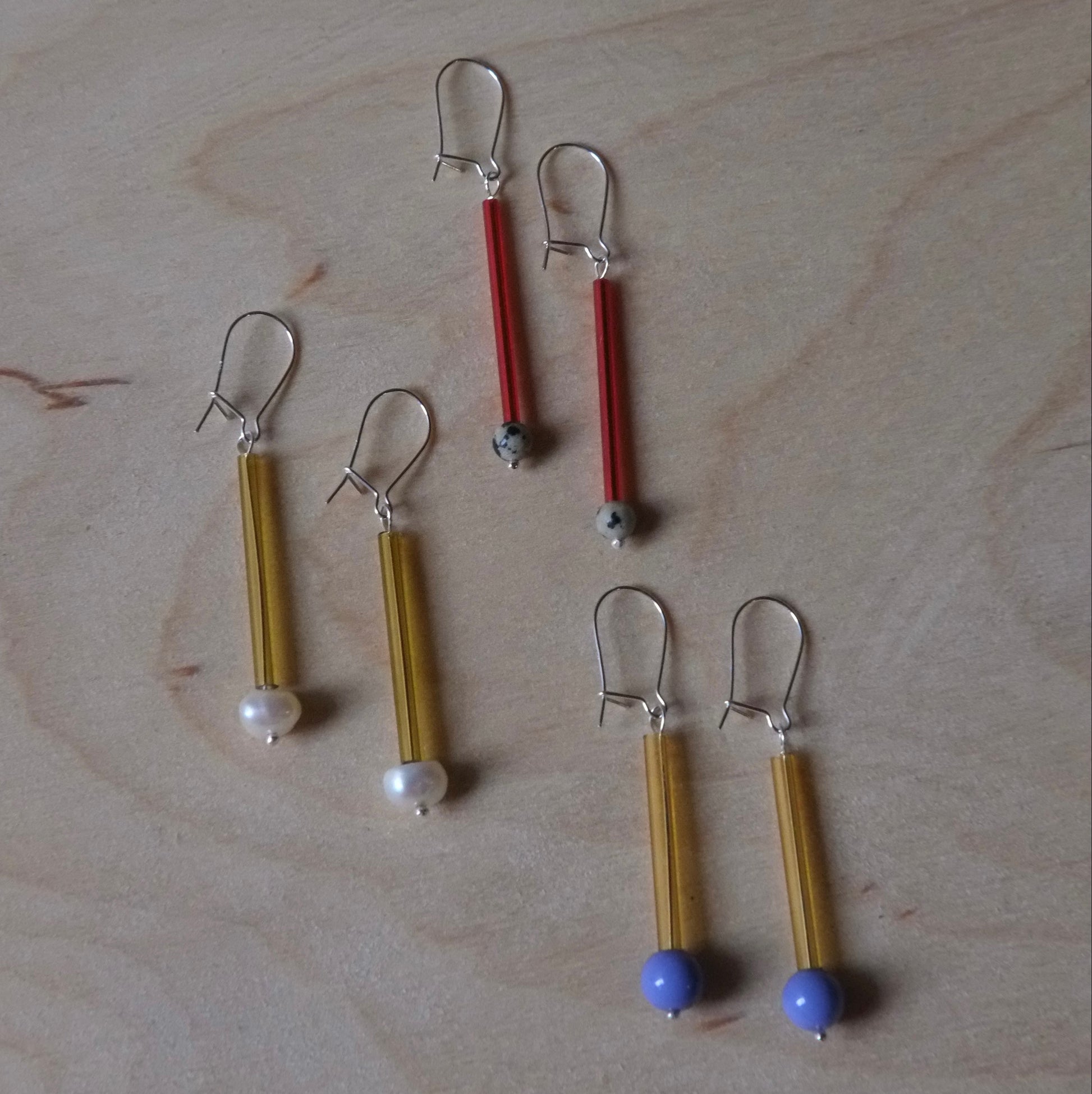 Five pairs of handmade earrings laid out on a light wood grain surface. The earrings feature gold-toned metal wire wrapped tubes, with one pair tipped in red, one in blue, and two with small white beads at the bottom.