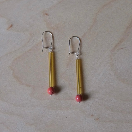 A pair of handmade dangle earrings featuring gold-toned tubes with small red beads at the bottom and silver fishhook backings, displayed on a light wood grain surface.