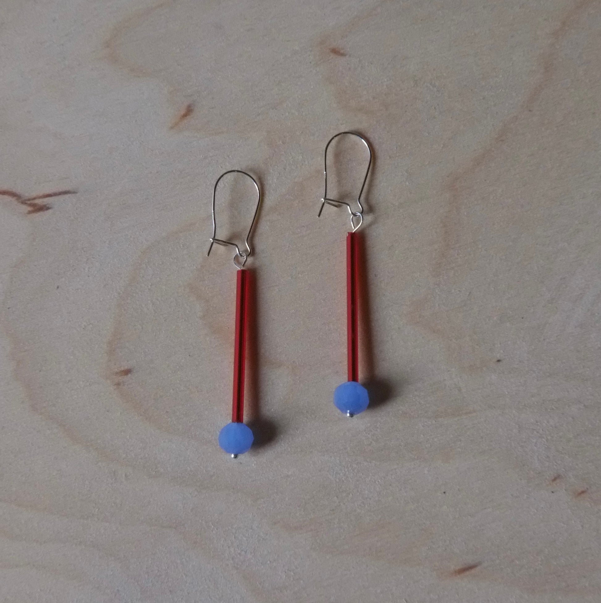 A pair of handmade dangle earrings with red cylindrical beads, small light blue round beads, and silver fishhook backings, displayed on a light wood grain surface.