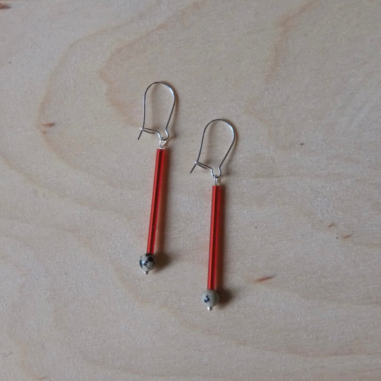 A pair of handmade dangle earrings with vibrant red glass rods, silver-toned metal hooks, and small grey beads at the bottom, displayed on a light wood grain surface.