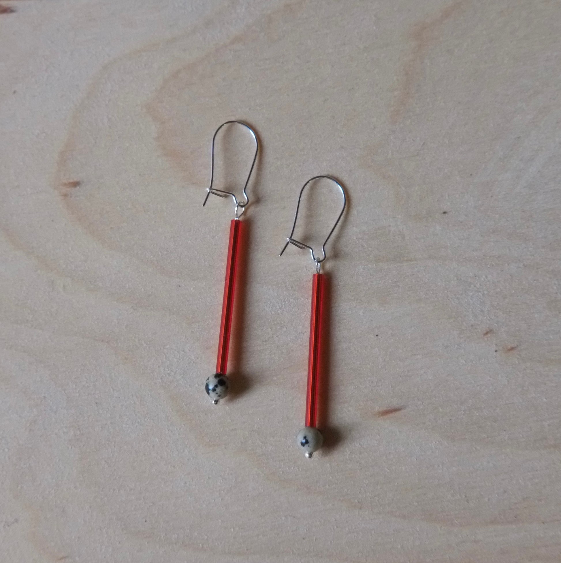 A pair of handmade dangle earrings with vibrant red glass rods, silver-toned metal hooks, and small grey beads at the bottom, displayed on a light wood grain surface.