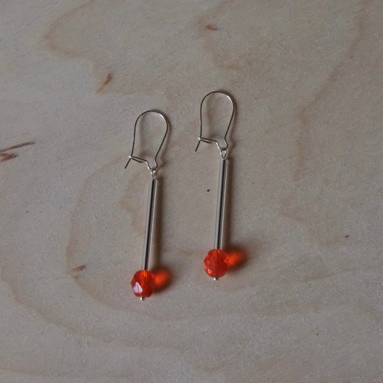 A pair of silver-toned drop earrings with small orange-red beads, displayed on a light wood grain surface.