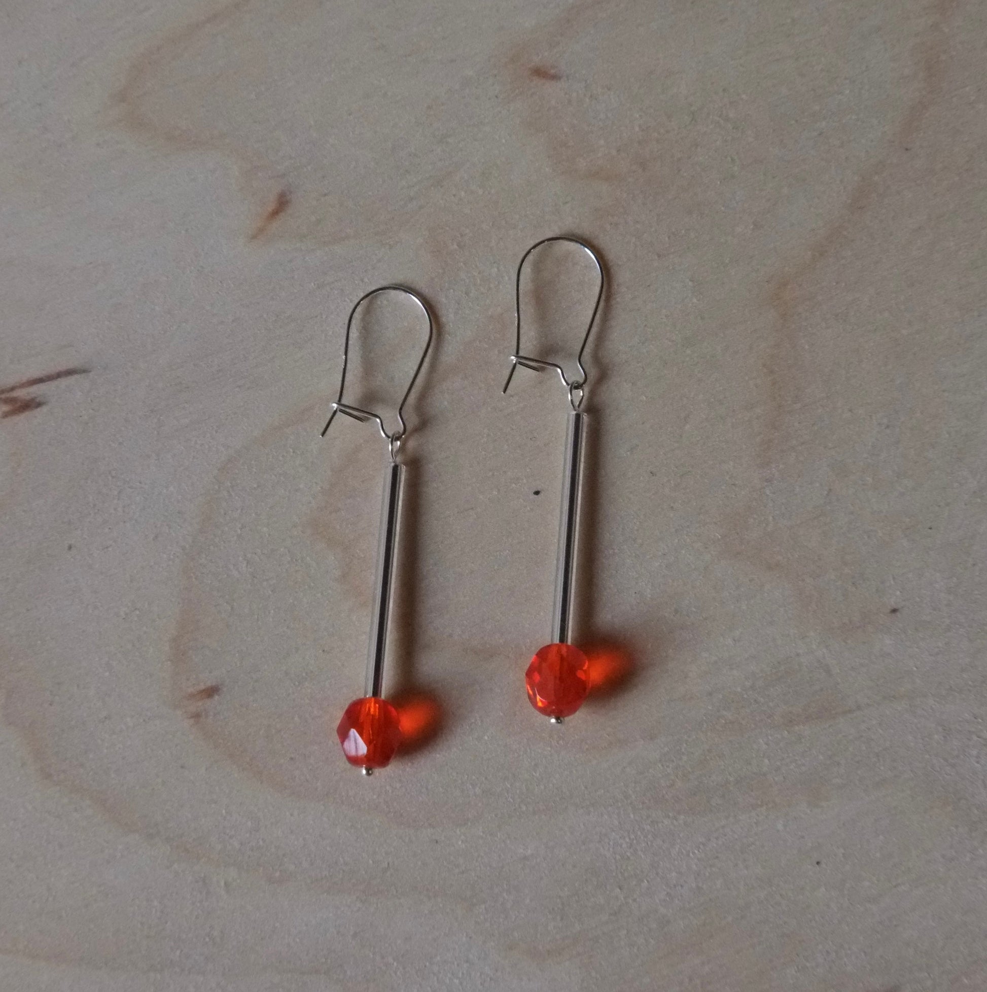A pair of silver-toned drop earrings with small orange-red beads, displayed on a light wood grain surface.