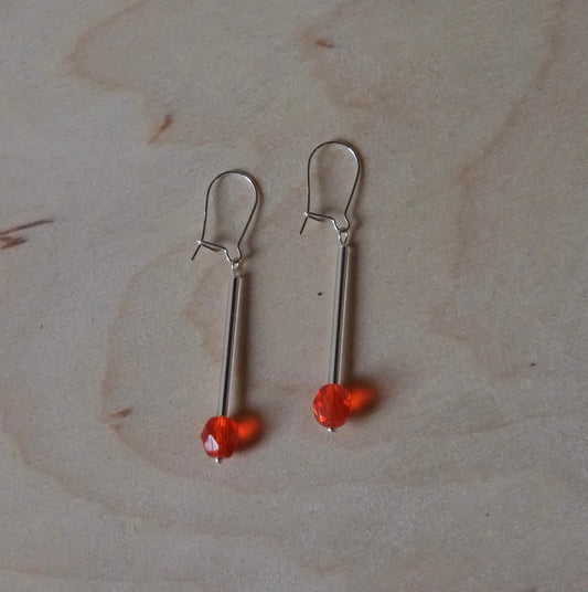 A pair of silver-toned drop earrings with small orange-red beads, displayed on a light wood grain surface.