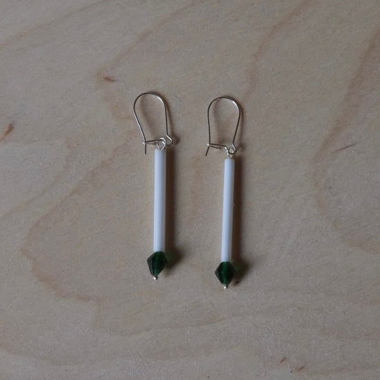 A pair of handmade dangle earrings with white cylindrical beads, dark green bead accents, and silver fishhook backings, displayed on a light wood grain surface.