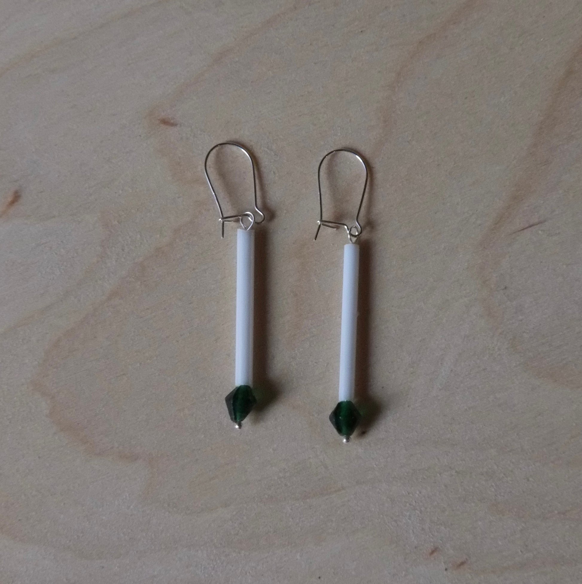 A pair of handmade dangle earrings with white cylindrical beads, dark green bead accents, and silver fishhook backings, displayed on a light wood grain surface.