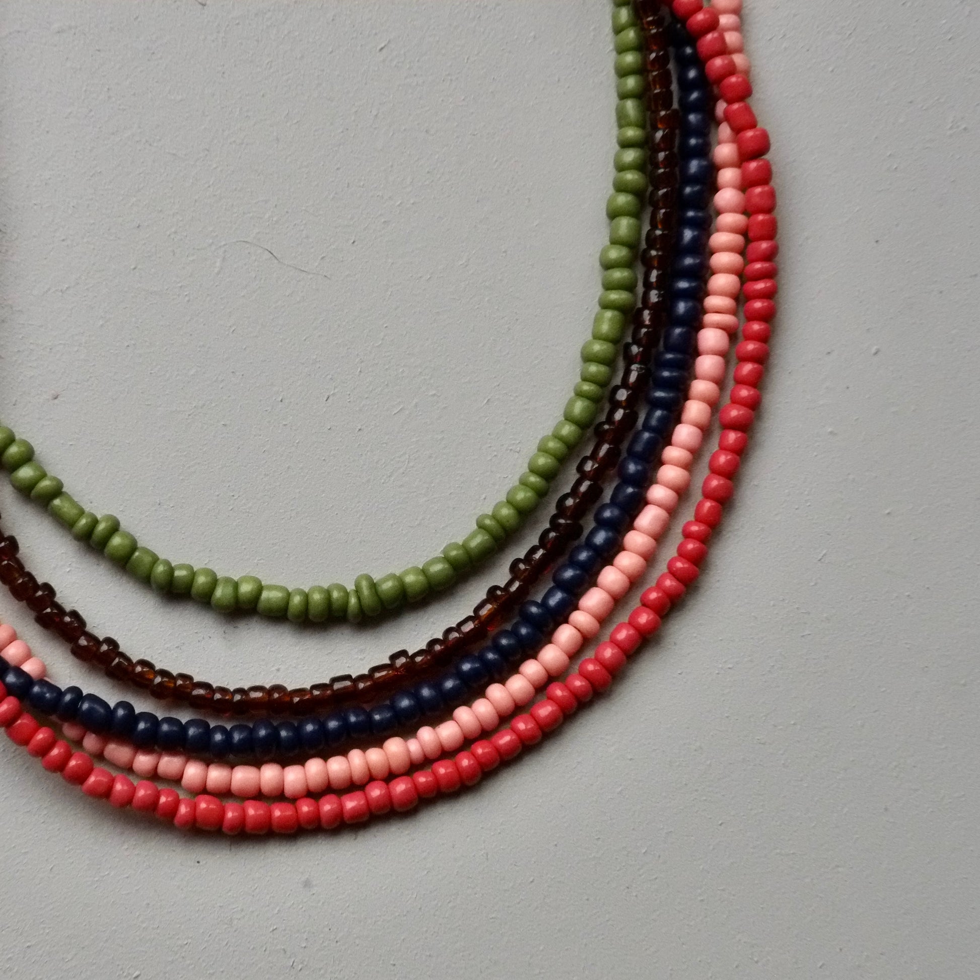 A set of five beaded strands arranged in a curved fan shape on a neutral gray background. The beads are small, cylindrical, and have a matte finish in various colors: olive green, dark brown, navy blue, light pink, and coral red.