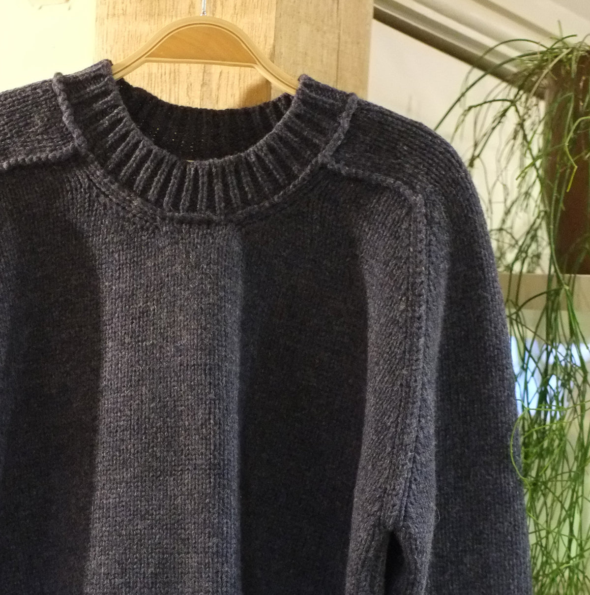 A navy knit sweater with a ribbed crew neck, dropped shoulders, and ribbed cuffs and hem, hanging on a wooden hanger against a rustic wooden display stand.
