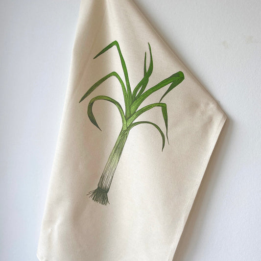 A beige cotton tea towel with a green illustration of a leek on it, hanging on a wall.