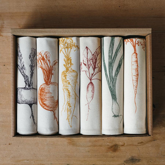 A set of six folded cloth napkins with vegetable illustrations, placed in a wooden box on a wooden surface.