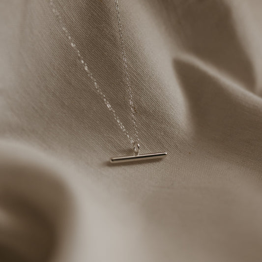 A silver bar necklace with a high polish pendant on a delicate trace chain, displayed on a textured background.